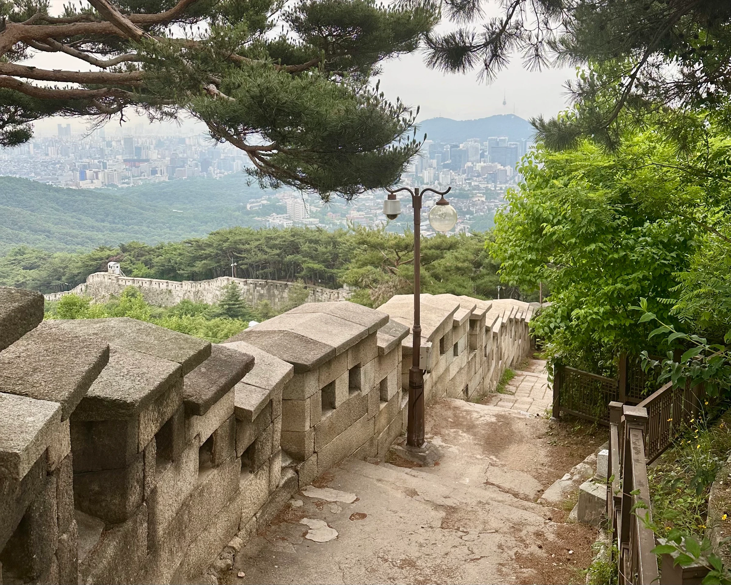 The crenellated stone fortress wall descending the ridge of Bugaksan among pine trees, with the hazy city and N Seoul Tower in the distance.