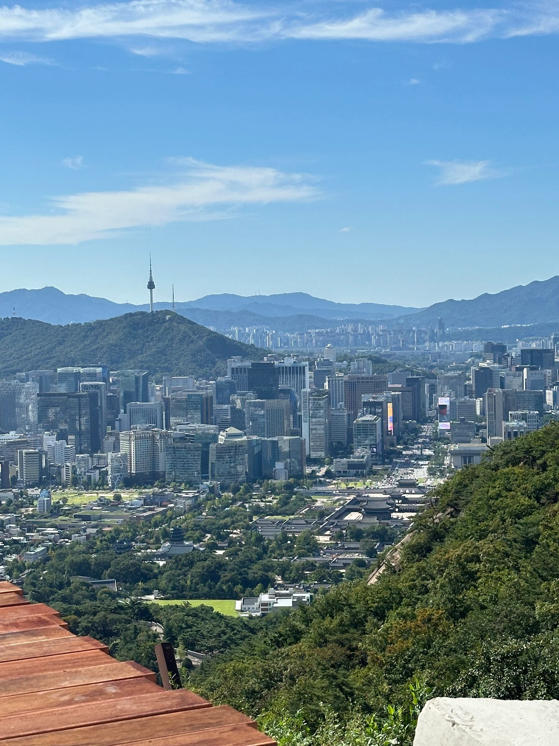 Looking south from the Bugaksan wall over the Blue House grounds and their lawn in the foreground to the tiled roofs of Gyeongbokgung and the line of Gwanghwamun Square, downtown towers behind and Namsan with N Seoul Tower on the skyline.