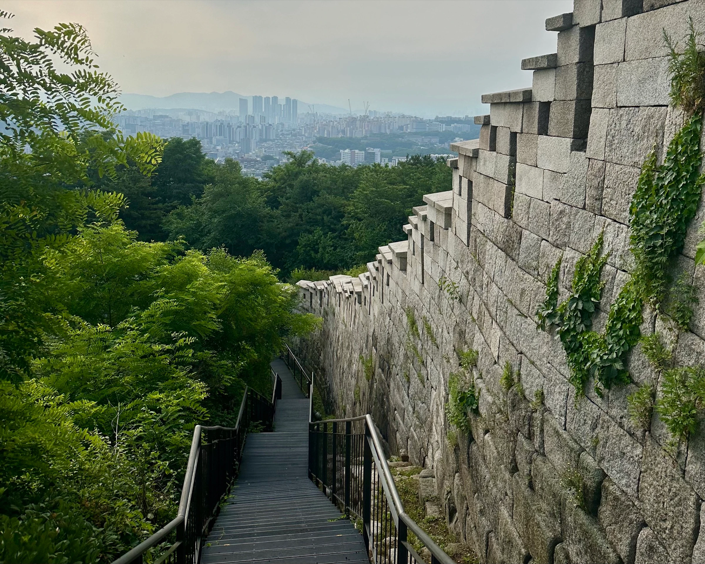 A steep wooden staircase descending alongside the towering granite blocks of the restored Seoul City Wall on Bugaksan, green forest to the left and the city beyond.
