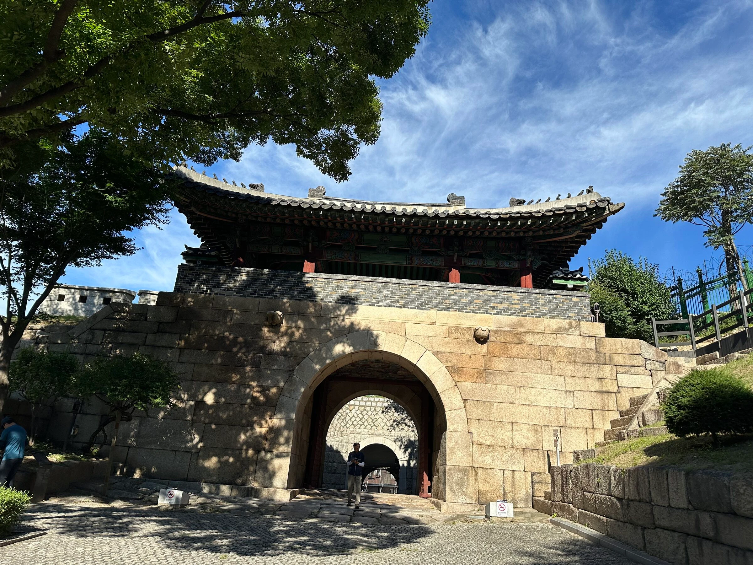 The arched stone gateway and painted pavilion of Changuimun under a bright sky, a large tree overhanging from the left and a round guard emplacement standing on the rampart beyond it.