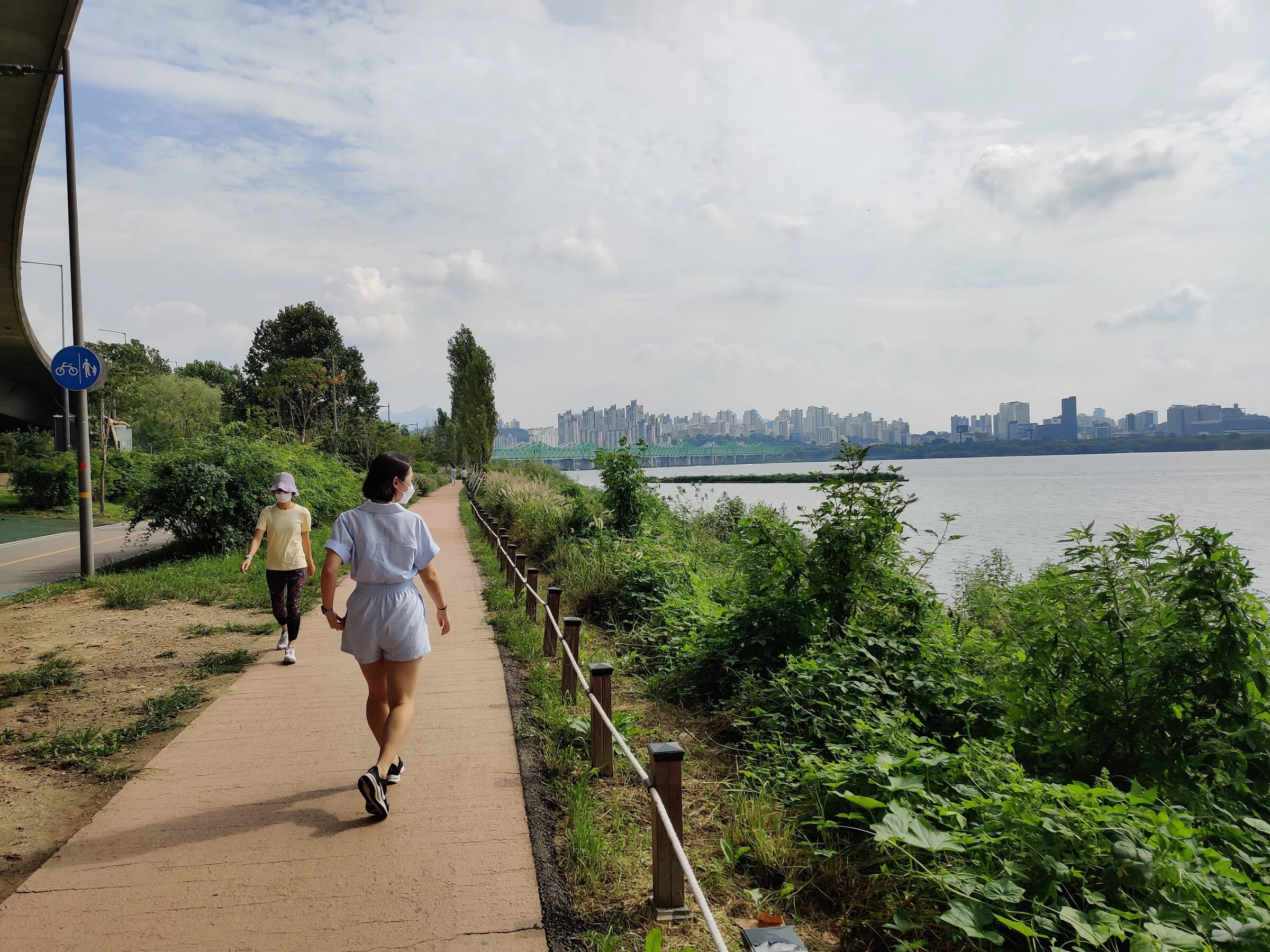Two people walking the Han River trail at Ichon on a summer afternoon, the river and the far bank behind them.