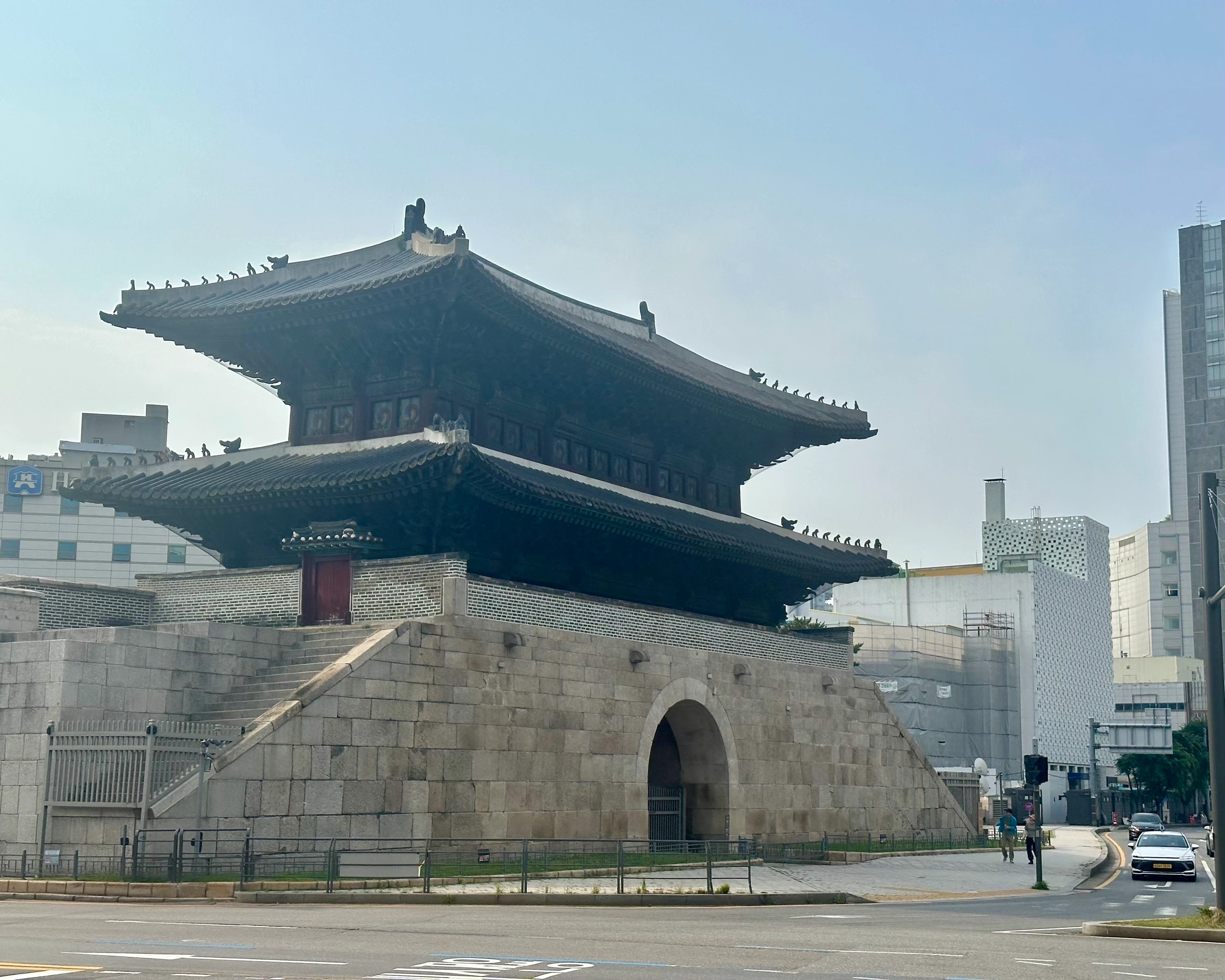 Heunginjimun, the Great East Gate (Dongdaemun), a two-tier wooden pavilion on a stone base with its arched gateway, pigeons lined along the eaves against a hazy sky.