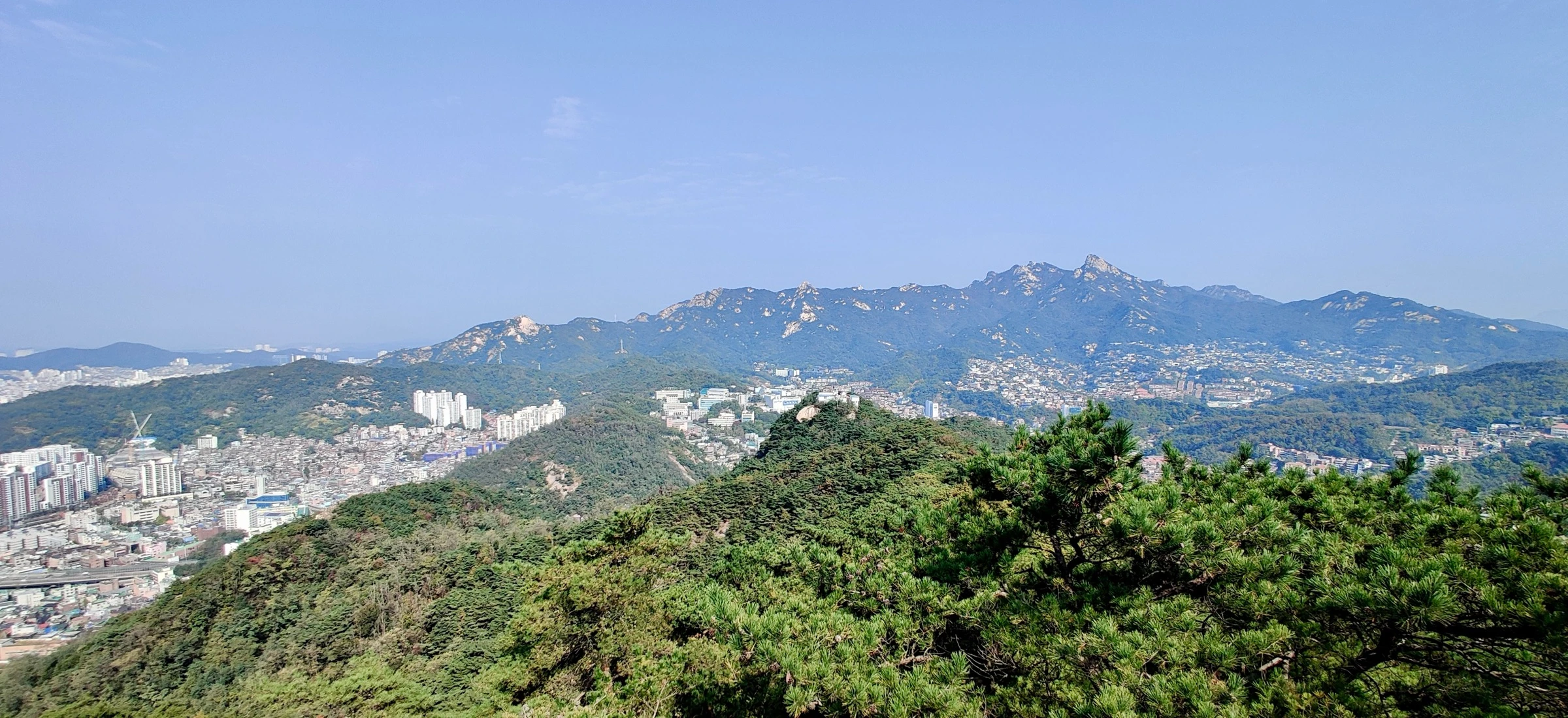 A wide panorama of the craggy granite peaks of the Inwangsan and Bugaksan range under a clear blue sky, the city visible below the forested ridgelines.