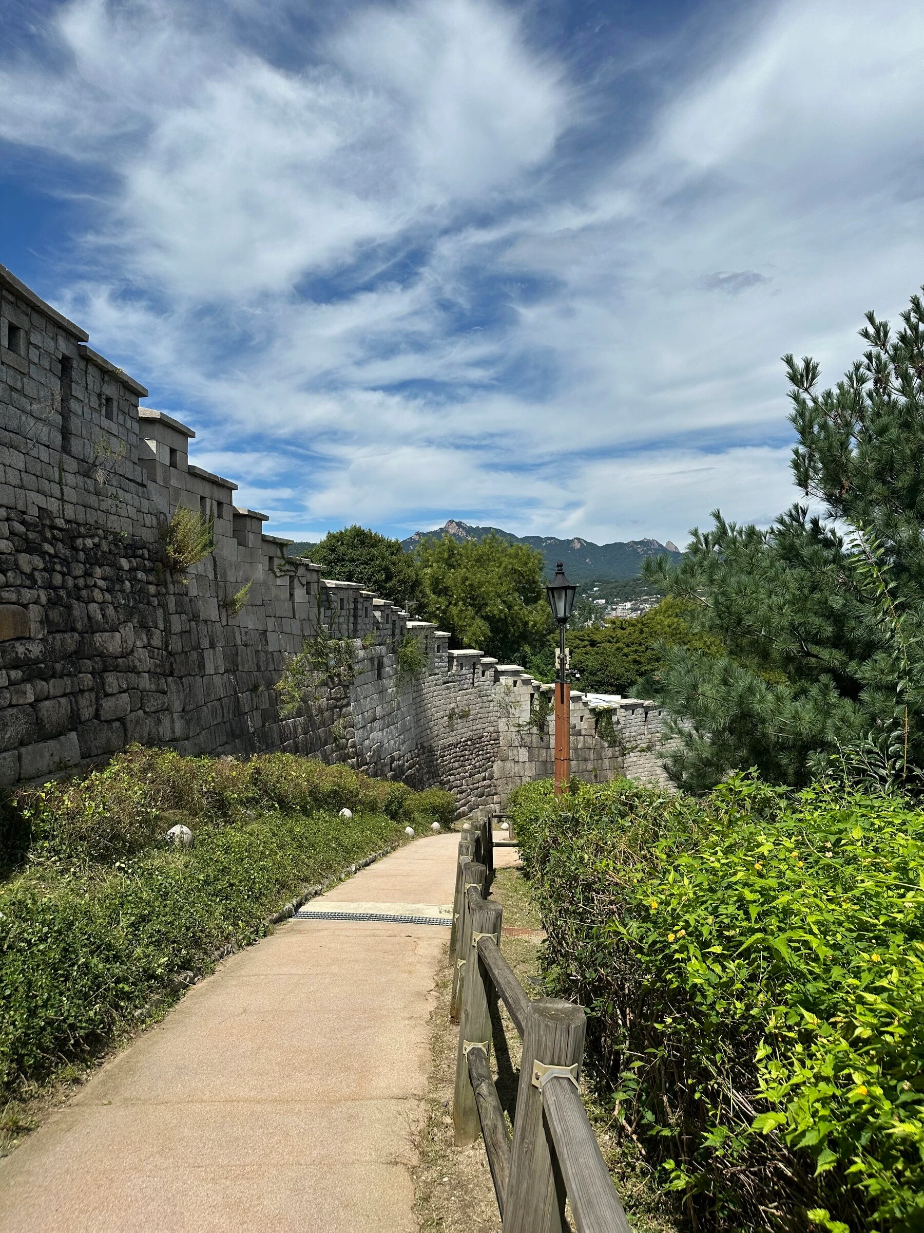 The clean paved path running along the base of the Naksan wall with nobody on it, a lamp post beside the trail and the Bukhansan ridge blue in the distance.