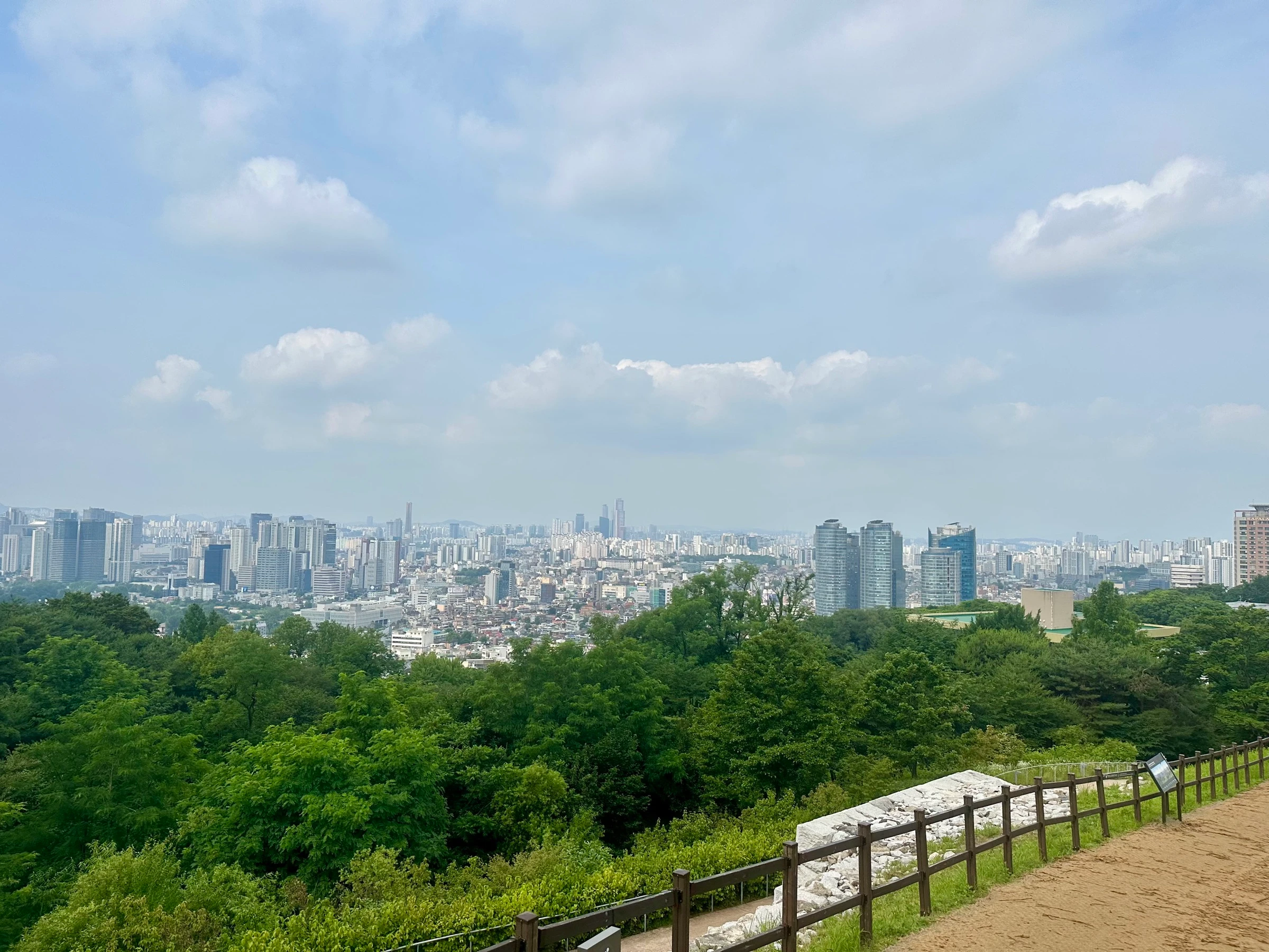 The southern Seoul skyline seen from the Namsan section of the city wall, green forest in front and tower clusters spreading to the horizon, with an excavated wall foundation and wooden fence in the foreground.
