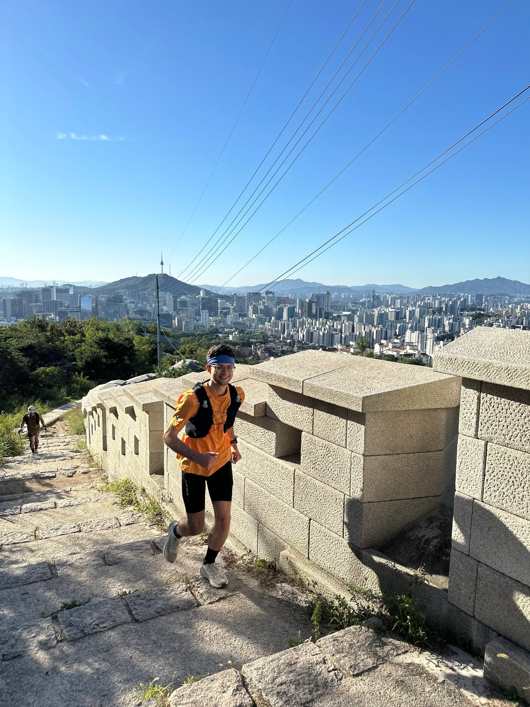 Quintin mid-stride and smiling on the wall path high on Inwangsan, the granite parapet beside him and the whole of central Seoul opening out behind.