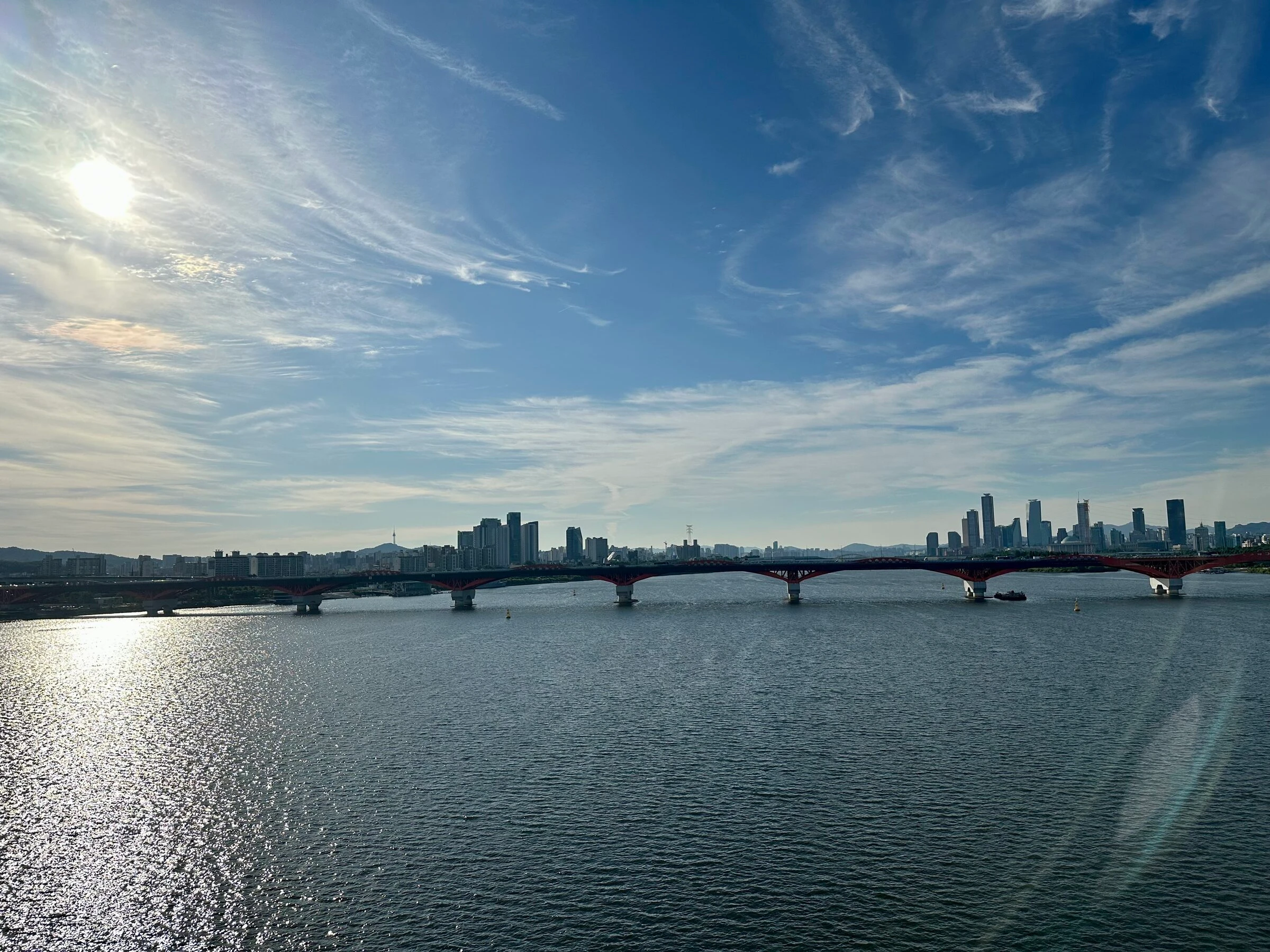 Seongsan Bridge silhouetted against a bright morning sky, sunlight scattering across the Han River with the Seoul skyline beyond.