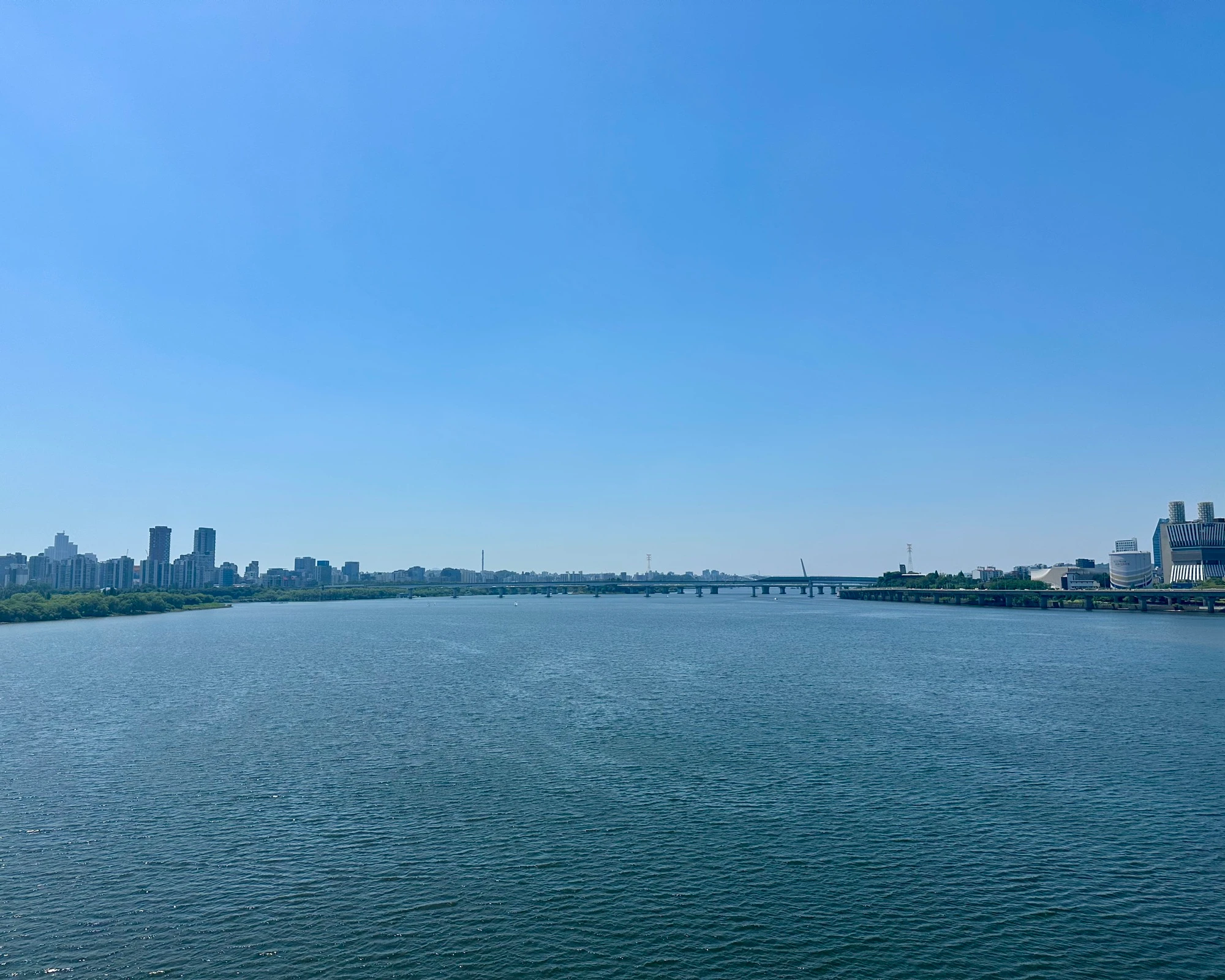 A wide open view of the Han River from a bridge on a clear blue-sky day, distant skyline and bridges along the water.